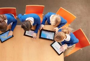 As an example of school tech, four kids sit at a large table, each working with a tablet. They are all wearing a uniform with a white shirt covered with a blue sweater. Photo courtesy of new-educ.com